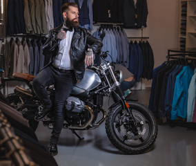 Fototapeta na wymiar Stylish tattooed bearded man with dressed in black leather jacket and bow tie posing near retro sports motorbike at men's clothing store.