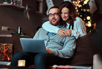 Obraz premium Portrait of an adorable couple with laptop - charming woman in Santa hat hugging her man and looking at camera.
