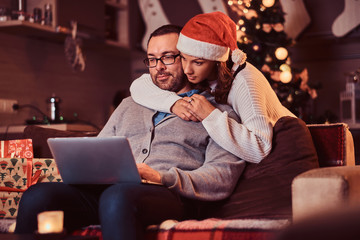 Portrait of an adorable couple - charming woman in Santa hat hugging her man and using a laptop.