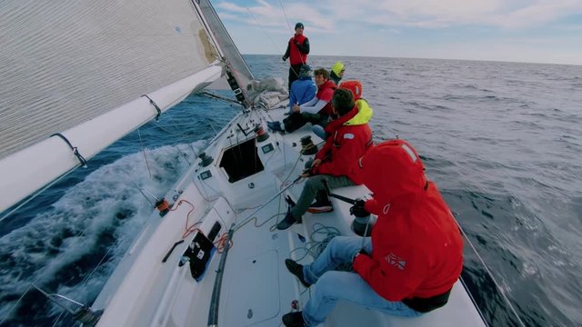 Group of men in red sailing outdoor jackets sit on deck of professional racing sailboat, sailors listen to captain give orders, sail in open waters of sea or ocean. Team work for common win or success