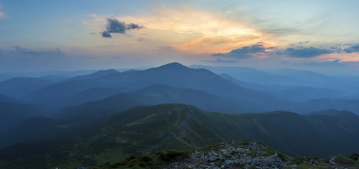 Fantastic sunrise or sunset over green mountain ridge covered with dense blue fog. Bright orange sun raising in  soft cloudy sky over distant horizon. Beauty of nature, tourism and traveling concept.