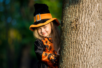 Obraz premium Little cute girl in witch costume peeking out from behind a tree in the park for the Halloween holiday.