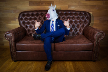 Funny unicorn in elegant suit sits on sofa like a boss and showing gesture thumbs up. Portrait of...