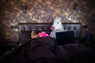 Blonde girl sleeping with funny man in comical mask. Unusual couple lying on the bed. Unicorn watching movie on laptop at the stylish bedroom.