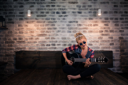 Glamour blonde girl in causal clothes playing music on guitar in stylish apartment. Unusual young woman sits on the bed at modern home.