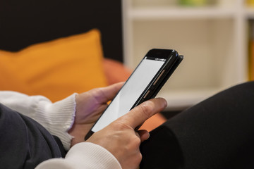 young woman looks at her smarthphone with white screen, sitting on the sofa, in front of the tv