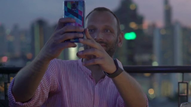 Young man with drink taking selfie with cellphone in skybar at night
