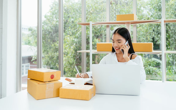 Young asian business owner woman communicate with customer through smart phone for order purchasing in modern home office with package boxes on table