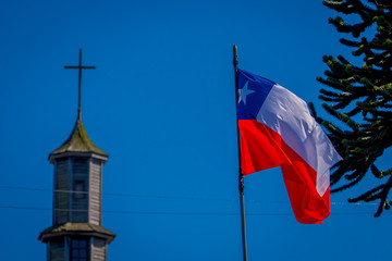 Exterior detail view of chilean flag waving with a blurred vilupulli church behind, one of world heritage wooden churches located at Chiloe island, Chile