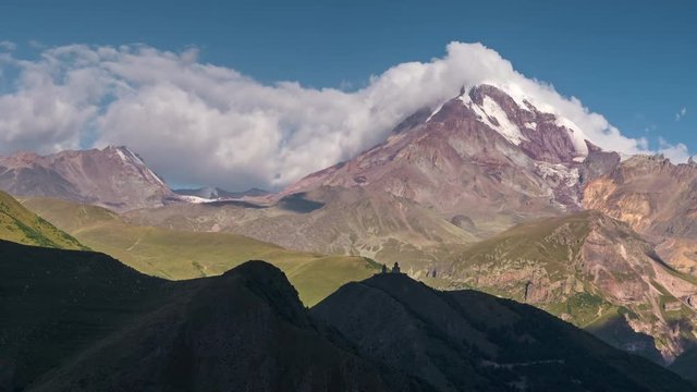 Caucasian highland. Old church and Kazbek mountain peak with snow cap and glacier in sun rays with moving shadows and rolling clouds time lapse