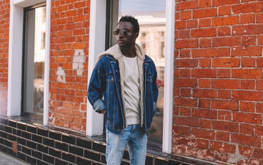 Fashion african man wearing jeans jacket poses on city street, brick wall background