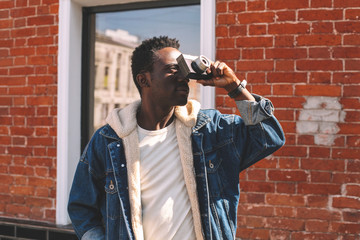 Fashion african man with vintage film camera taking picture on city street, brick wall background