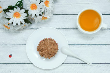 A cupcake on a white plate, a cup of green tea, a bunch of daisy flowers. White wooden background.