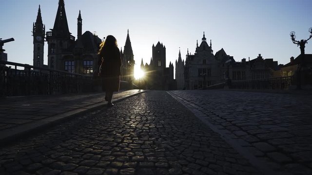 Cobblestone pavement on St. Michael Bridge in Ghent.Following young girl beck view silhouette in back light at background belfry towers and roofs ancient buildings