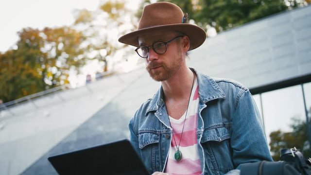 Portrait of young bearded hipster man and in hat sits on bench in city center in park using laptop, slow motion, close up