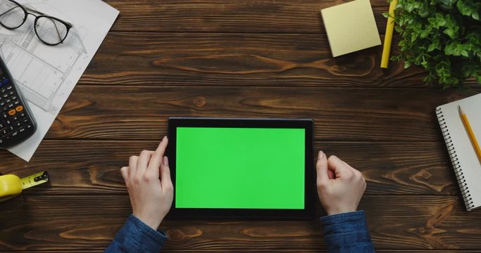 Top view on the black horizontal tablet computer on the wooden table of architect and Caucasian female hands scrolling and tapping on the green screen. Chroma key.
