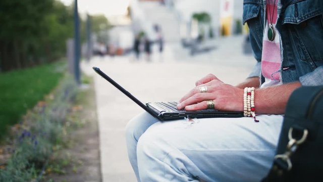 Close up shot of young hipster man and in hat sits on bench in city center in park using laptop, slow motion