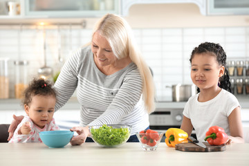 Female mature nanny feeding little African-American girl in kitchen
