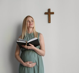 Young pregnant woman with Bible praying near light wall. Space for text