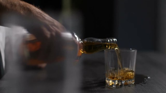 Closeup of wasted alcoholic man with shaking hands trying to refill glass with whiskey and spilling alcohol beverage on the dirty table. Adult man in alcohol abuse pouring whiskey into glass indoors.