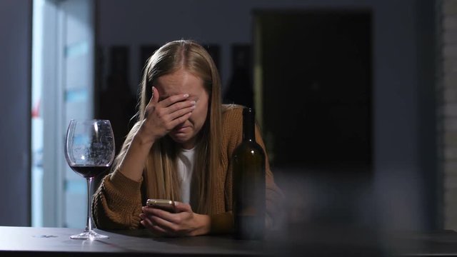 Rejected crying adult woman shocked by text message received on cellphone with glass and bottle of wine on the table. Depressed sad woman in tears checking phone feeling rejected after breakup message
