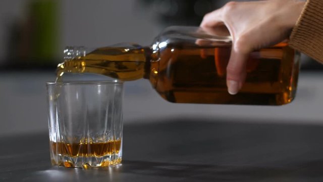 Closeup of female trembling hand pouring alcohol beverage into drinking glass and spilling alcohol on the table. Boozer woman refilling glass with whiskey, feeling dizzy after too much alcohol.