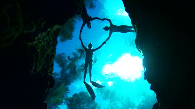 Scuba divers swim over cave holding hands, slow motion