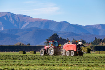 A red tractor and a baler working in a rural field in New Zealand making hay in the springtime