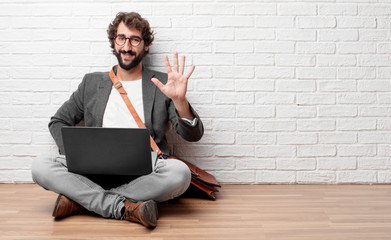 young man sitting on the floor smiling and looking satisfied and happy, counting number five with...