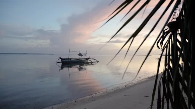 Traditional filipino bangka boat on calm waters at dusk as seen from beach behind palm tree