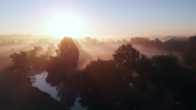 Aerial footage of a river and surrounding countryside at sunrise with low lying mist and a clear sky with birds flying past the camera.