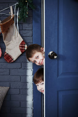 Two boys peeking out from behind the open door in the gray room of the Christmas decoration.