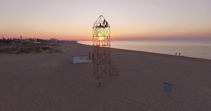 Dawn on the beach. We see the sun through the openwork tower, in which several people meet dawn. The camera is gradually rising along the tower.