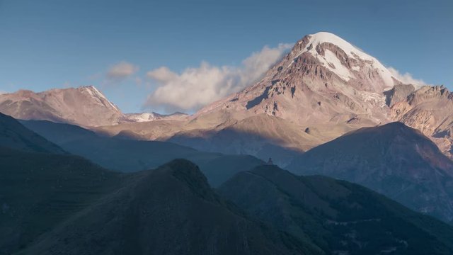 Sunrise in highlands. Kazbek mountain peak with snow cap and glacier in sun rays with moving shadows and clouds time lapse