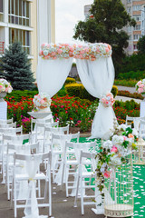 Fototapeta na wymiar Beautiful decoration of the wedding ceremony. White wooden chairs for guests on either side of the walkway, which leads to festive arch decorated with flowers