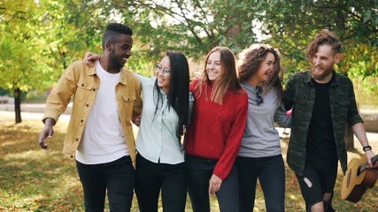 Slow motion of happy young people friends walking together in park in autumn holding guitar talking and laughing. Youth lifestyle, culture and people concept.