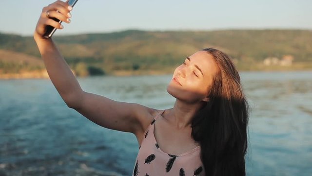 A half-body view of a beautiful girl with long loose hair making a photo of herself in front of the river with mountains at sunset