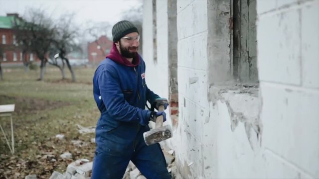 A side view of a worker smashing a wall with a sledgehammer. Some parts of the building are already destroyed. The holes are square shaped. The bricks on the ground.