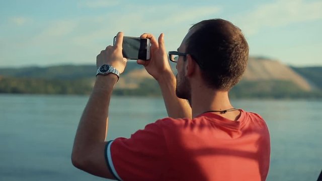 Back half-body view of a young man making a video on his smartphone of the river and mountains