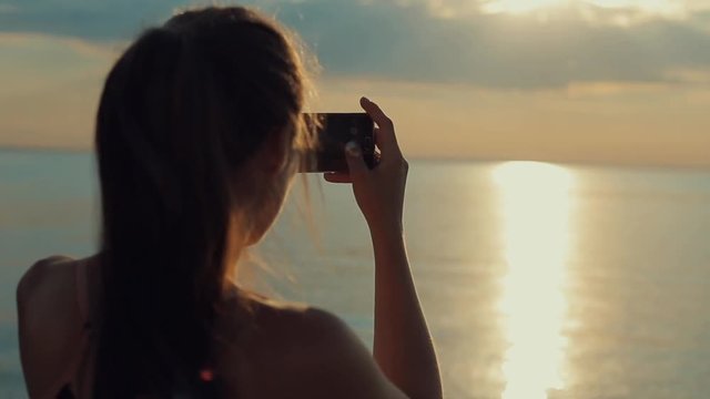 Back view of a beautiful girl taking a picture of the river at sunset. Taking photos on smartphone