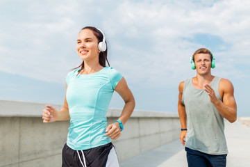 sport, people and technology concept - happy couple with headphones and fitness trackers running outdoors
