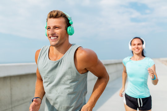 Sport, People And Technology Concept - Happy Couple With Headphones And Fitness Trackers Running Outdoors