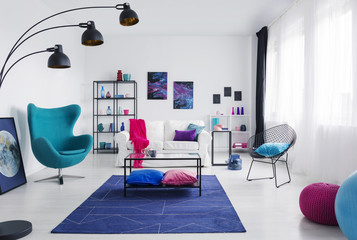 Table on blue carpet next to armchair in modern living room interior with posters and lamp. Real photo