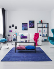 Posters on white wall above couch with pink blanket in flat interior with table on blue carpet. Real photo