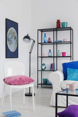 Lamp next to white chair with pink pillow in living room interior with poster and sofa. Real photo