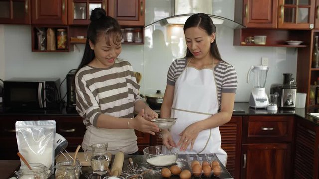 Attractive Asian woman and her young beautiful daughter sieving flour for cookie dough and smiling happily while standing in home kitchen