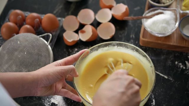 Close-up view of female hands mixing cookie dough with whisk on messy  kitchen table with eggshells and baking ingredients