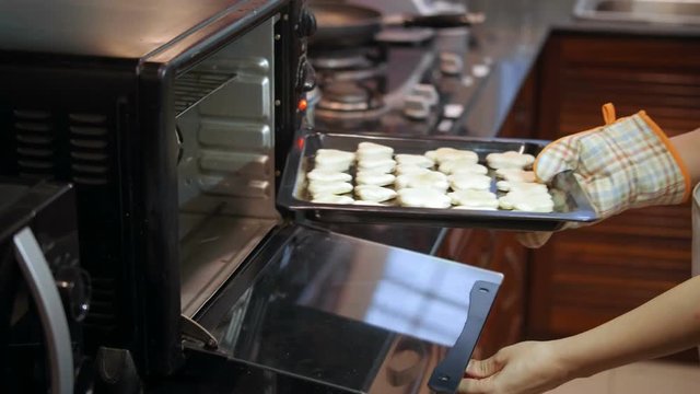 Unrecognizable woman wearing oven glove putting baking sheet with cut out cookies into preheated oven in domestic kitchen