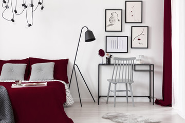 A gallery of illustrations on a white wall above a small desk which is next to a black metal lamp and a burgundy bed in a modern bedroom interior. Real photo.