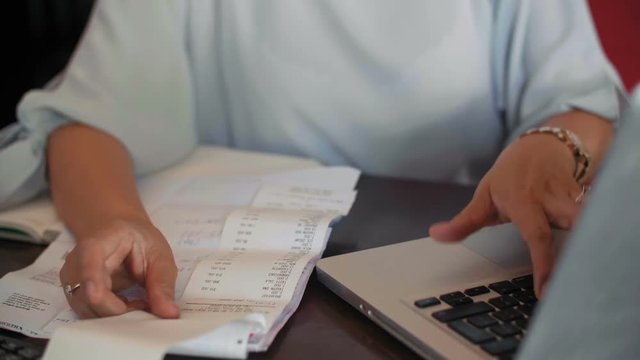Tilt-up shot of young Asian businesswoman calculating bar expenses on laptop while sitting at table with papers and receipts